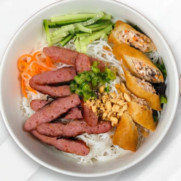A white bowl of vermicelli noodles with sliced sausage, spring rolls, cucumbers, carrots, and peanuts.